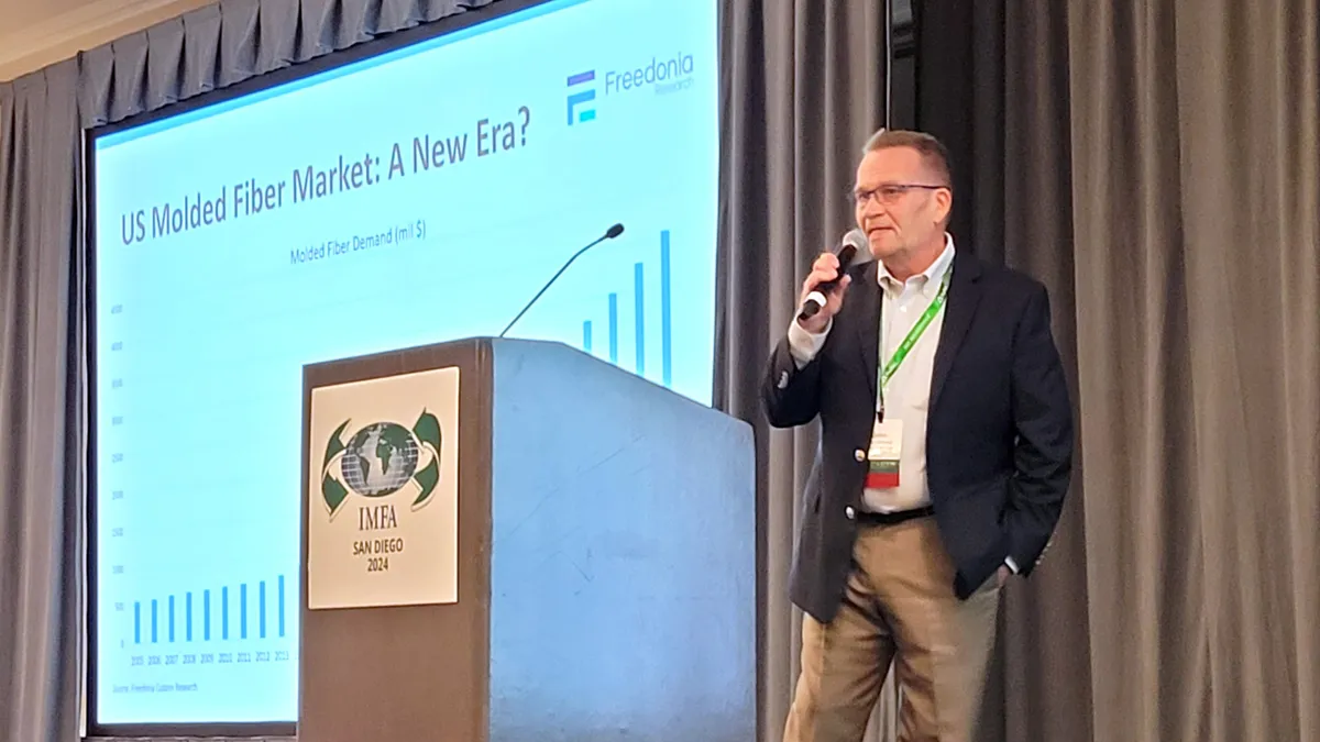 A person speaks into a microphone from a stage where they are next to a podium with a placard branded for the IMFA 2024 conference.