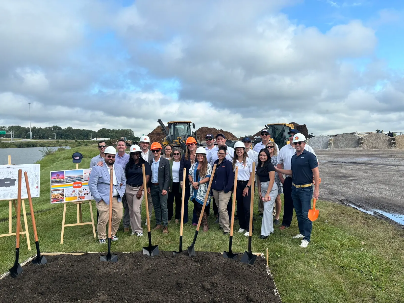 A photo of a number of people standing begind a patch of bare earth with shovels stuck in it in an open field. There is construction equipment behind them. A sign on an easel next to them shows several small pictures of a retail locations. Text in the center fo the sign says "Wally's."
