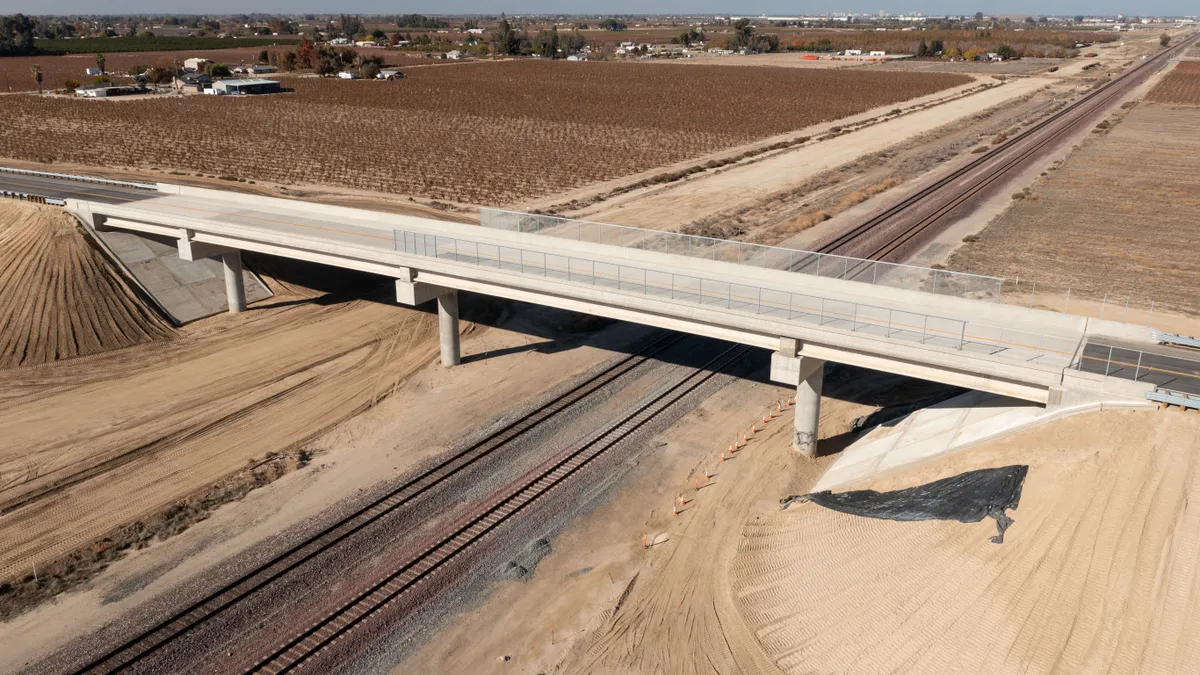 Two railroad tracks pass under a white concrete bridge with a farm field in the background.