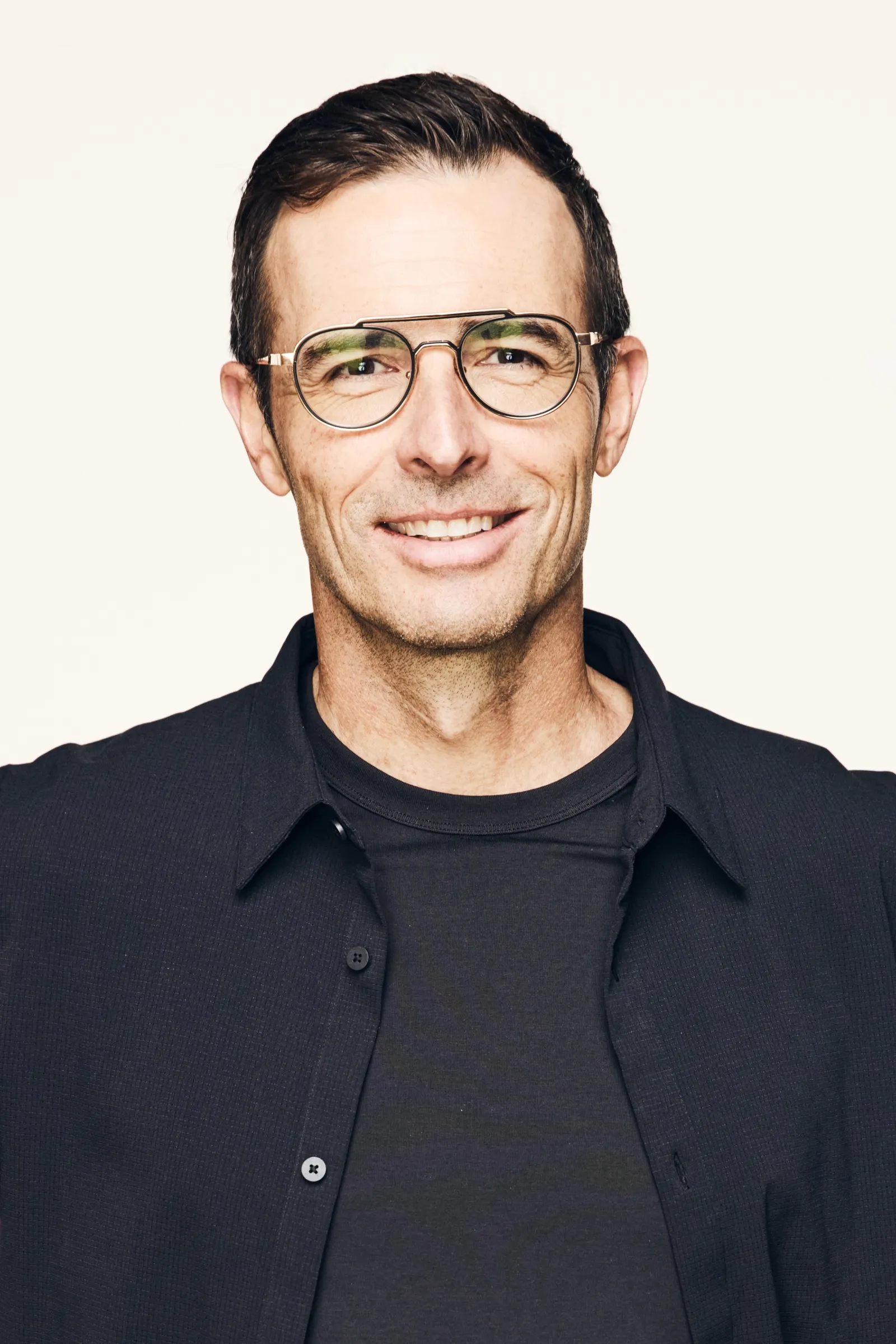 A person with short hair and glasses, wearing a dark shirt, smiles into the camera.