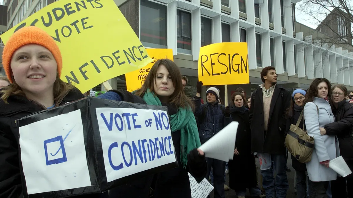 Crowd of protesters stand in front of building with signs saying "Vote of no confidence" and "Resign."