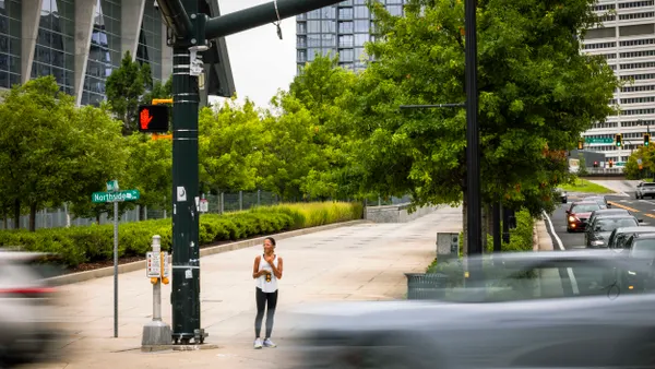 A person stands on a sidewalk next to a traffic signal as cars whiz by.
