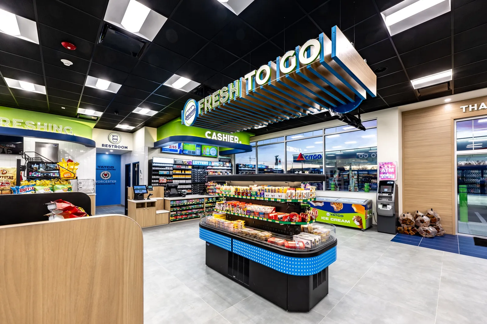 A photo of the interior of a store, showing the front of the store, including the checkout aera and a standalone cooler with grab-and-go items. The store is decorated in greens and blues along with wood tones.