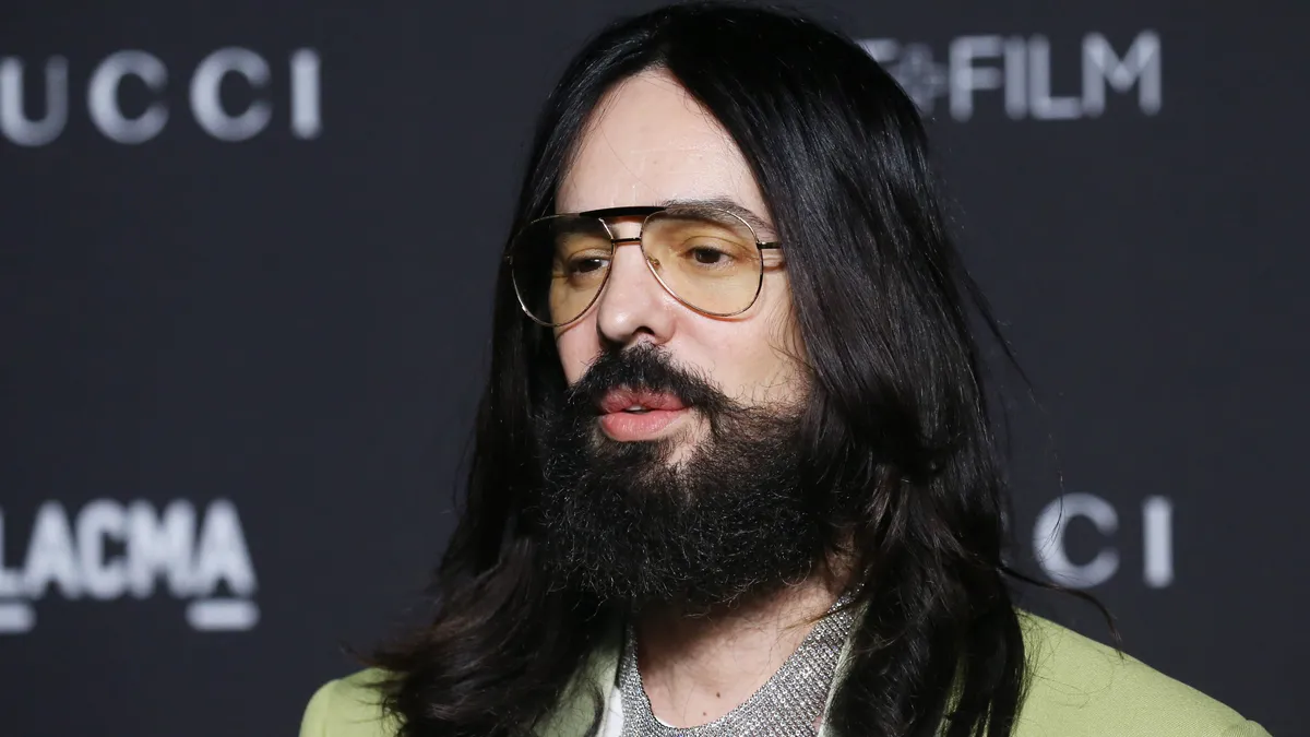 Alessandro Michele, with shoulder-length brown hair, wears a silver chain mail shirt under a gold brocade jacket and lightly tinted aviator sunglasses.