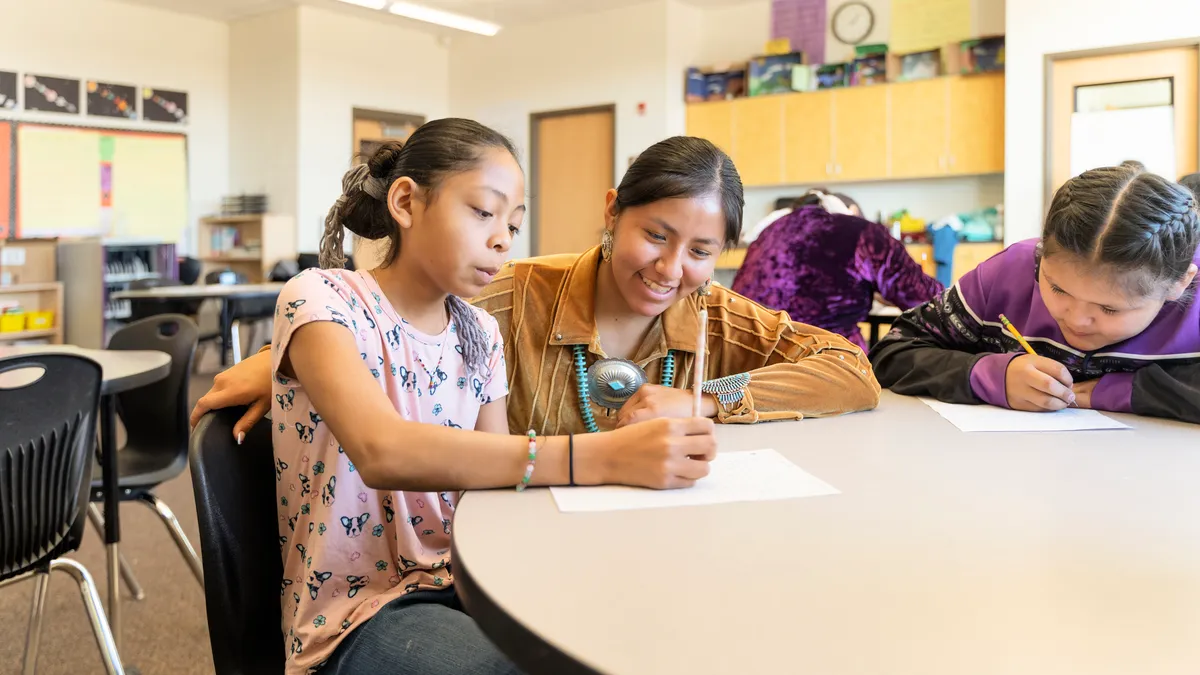 An adult is with two students at a desk in a classroom. The students are writing and reading.