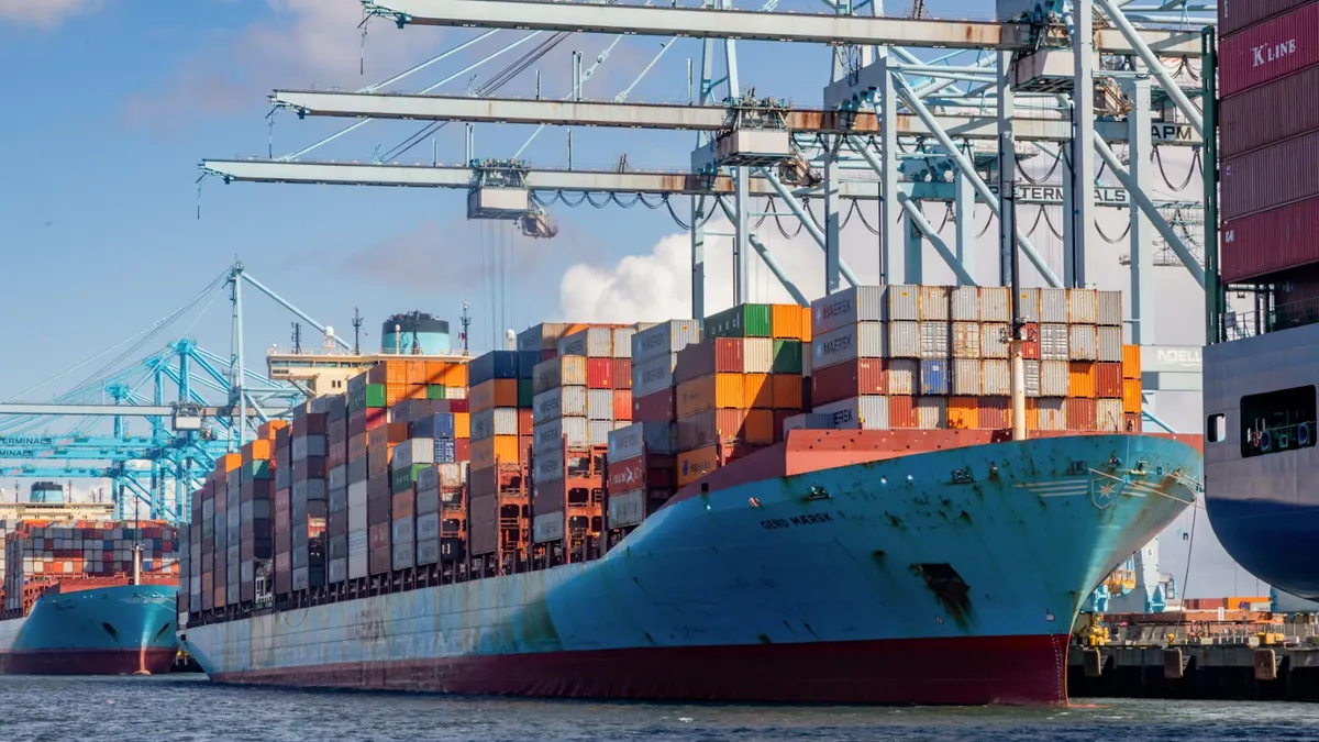 Dozens of multi-colored containers are stacked on a blue cargo ship docked at a port under cranes. The cargo ship is between two other vessels.