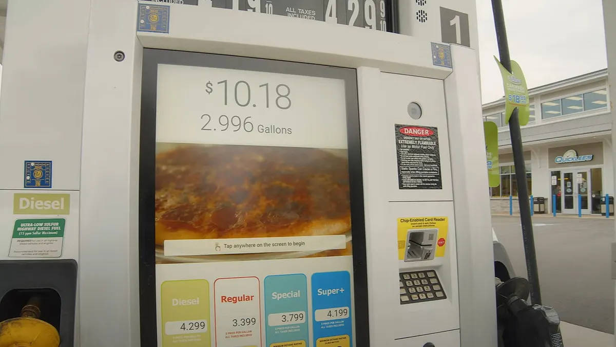 A screenshot showing a fuel pump with a large video screen. Text on the screen says $10.18. 2.996 gallons. Tap anywhere on the screen to begin.