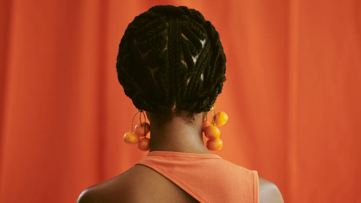 A Black feminine person with braids sits is faced away from the camera; they are wearing an ornate braided bob