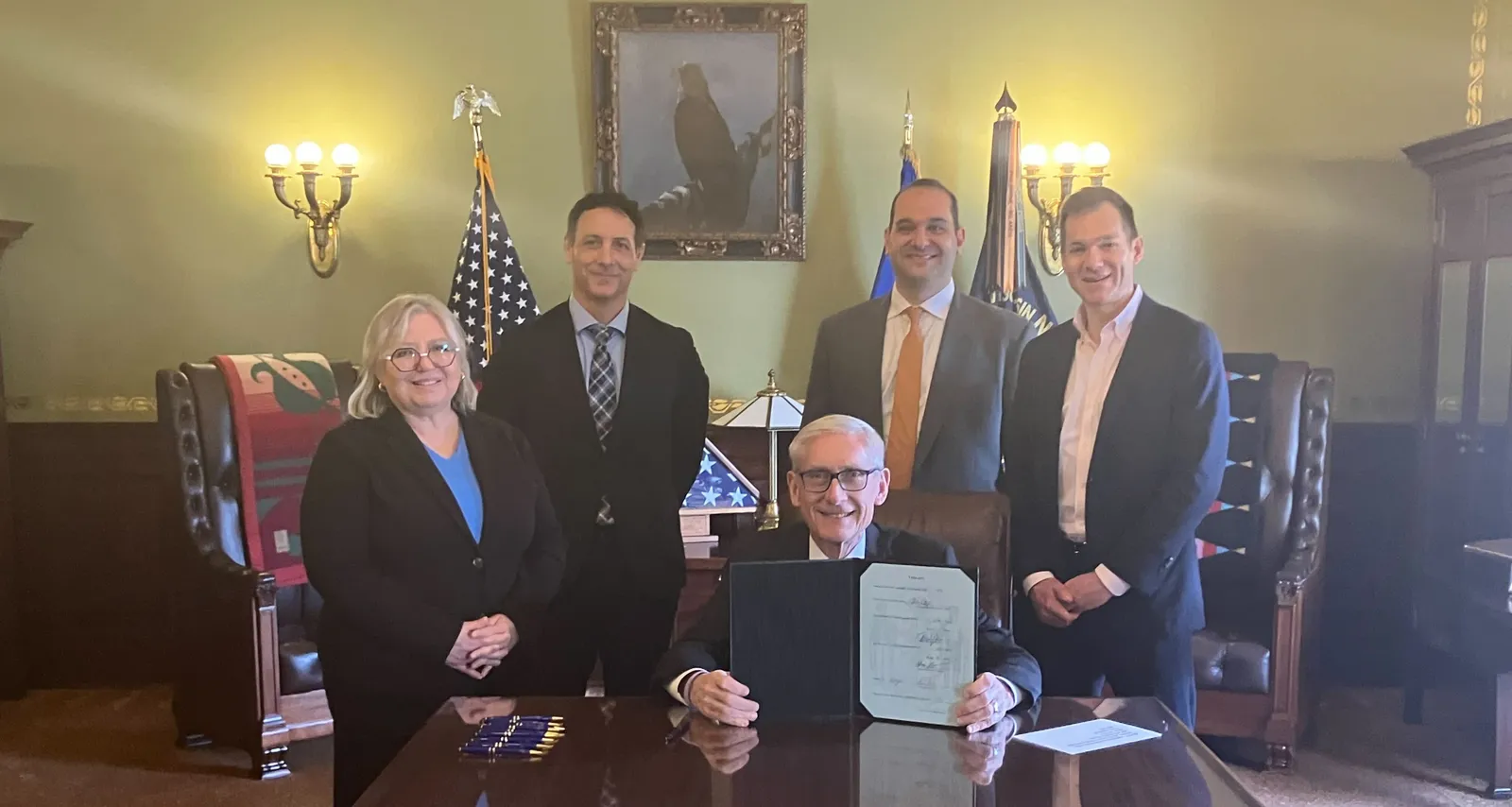 Governor Tony Evers sits at table holding up newly signed bill, with others standing behind him.