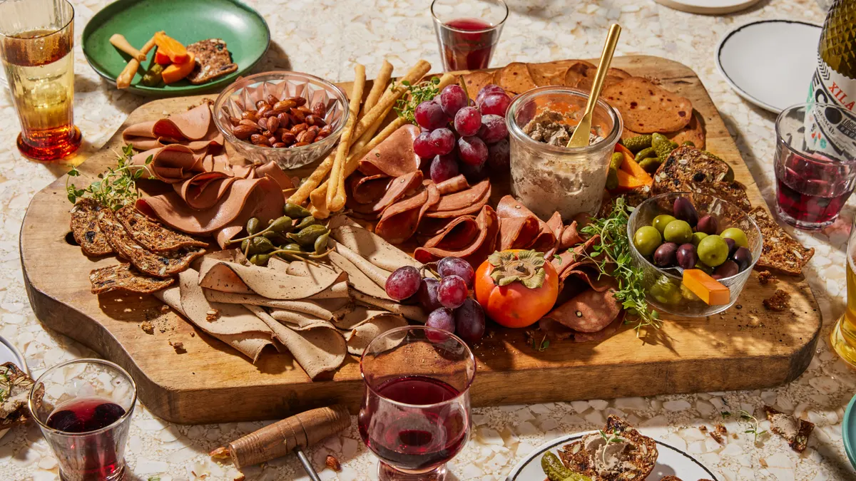 A variety of fermentation-made deli slices lie on a wooden charcuterie board, which also features grapes, breadsticks, a dish of olives, nuts and a dip. The board is on a table with plates and glasses