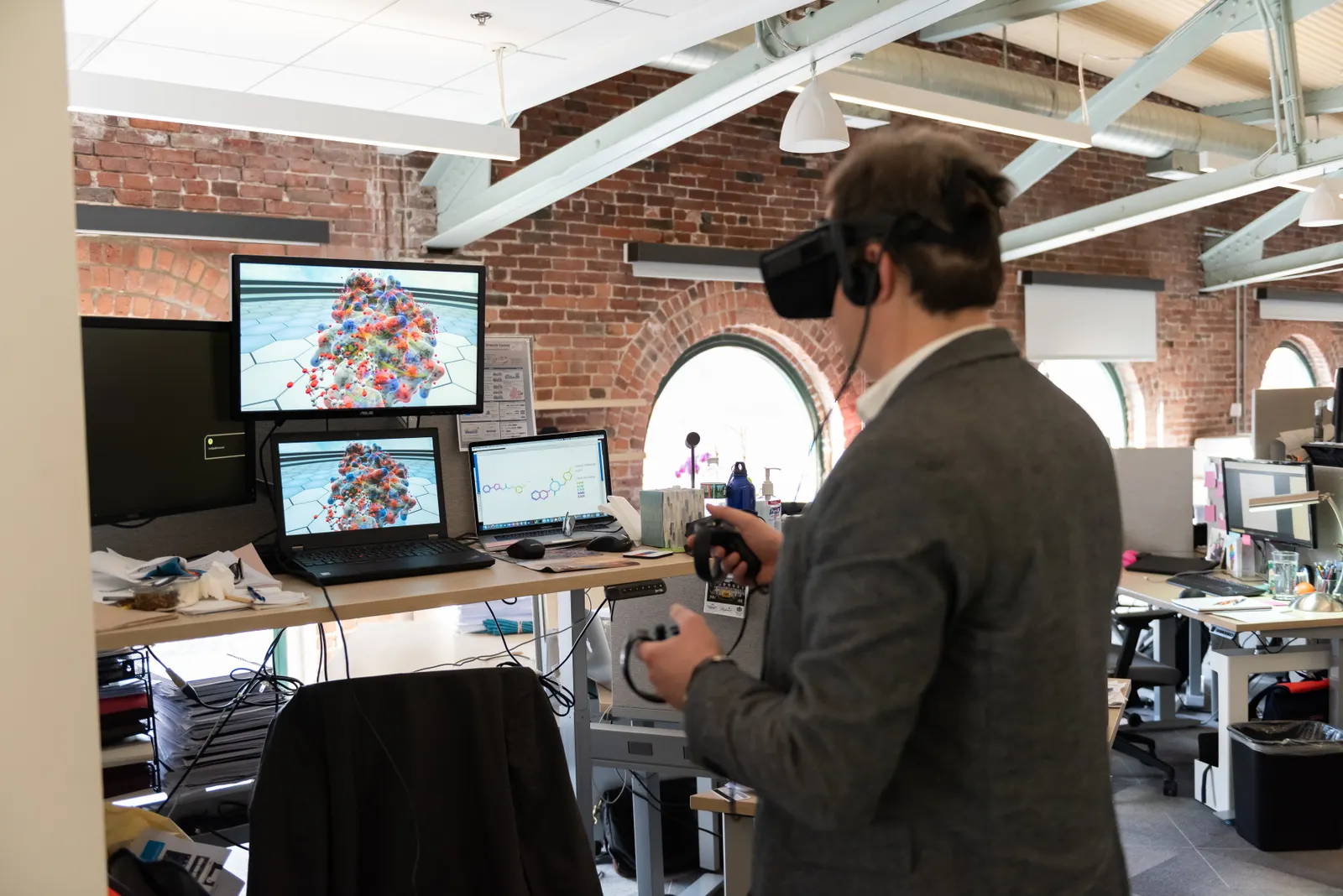 A person in an office uses a virtual reality headset to examine a 3D model on a computer screen.