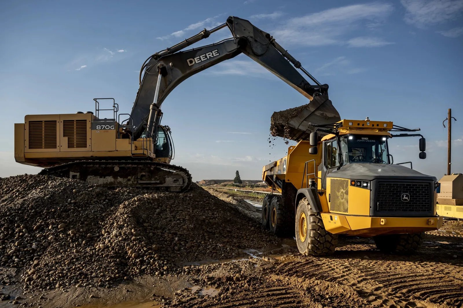 Two large industrial machines on dirt-covered ground. One pours dirt into the bucket of the large truck.