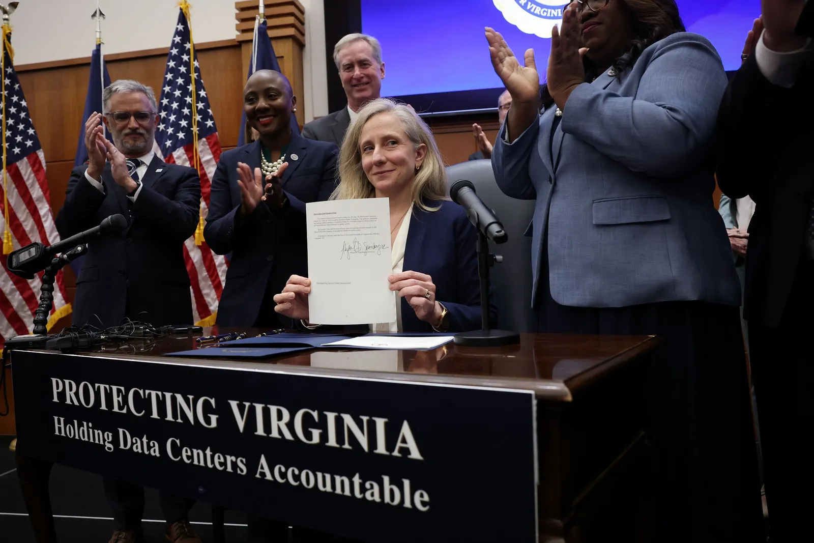 A person sitting at a table with a sign reading "Protect Virginia Holding Data Center Accountable" holds up a signed paper.