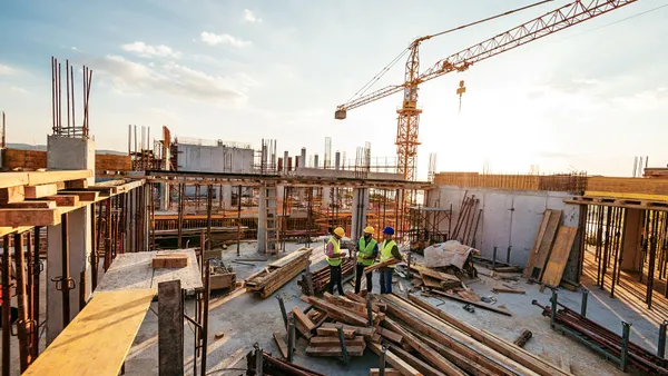 Three construction workers stand on a roof of a building under construction, surrounded by cranes and materials.