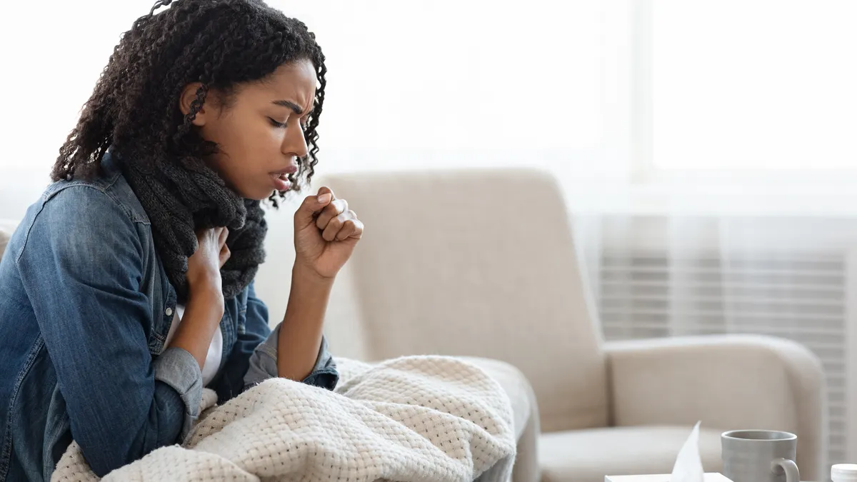 A woman coughs into her hand while sitting under a blanket.