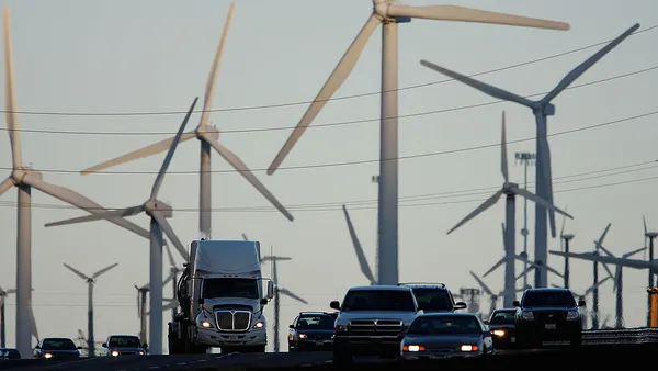 Diesel trucks and cars pass wind turbines along a freeway.