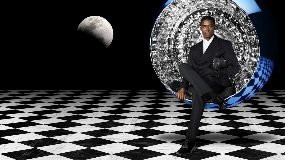 Damson Idris wears a black suit and sits in a silver lined circular seat holding a Prada bag.