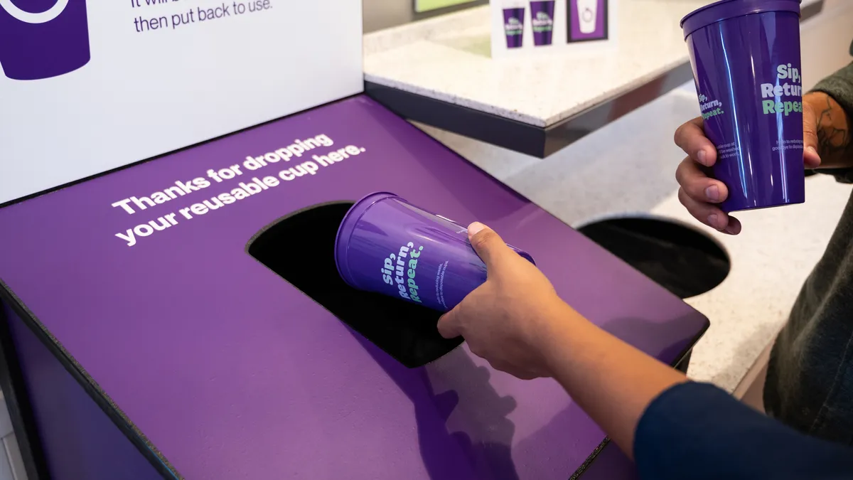 Two hands place two purple plastic reusable cups toward a receptacle that says "Thanks for dropping your reusable cup here."