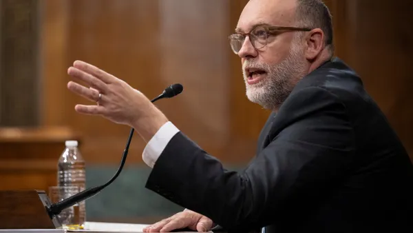 A person wearing a suit and tie speaks into a microphone while gesturing with their hand.