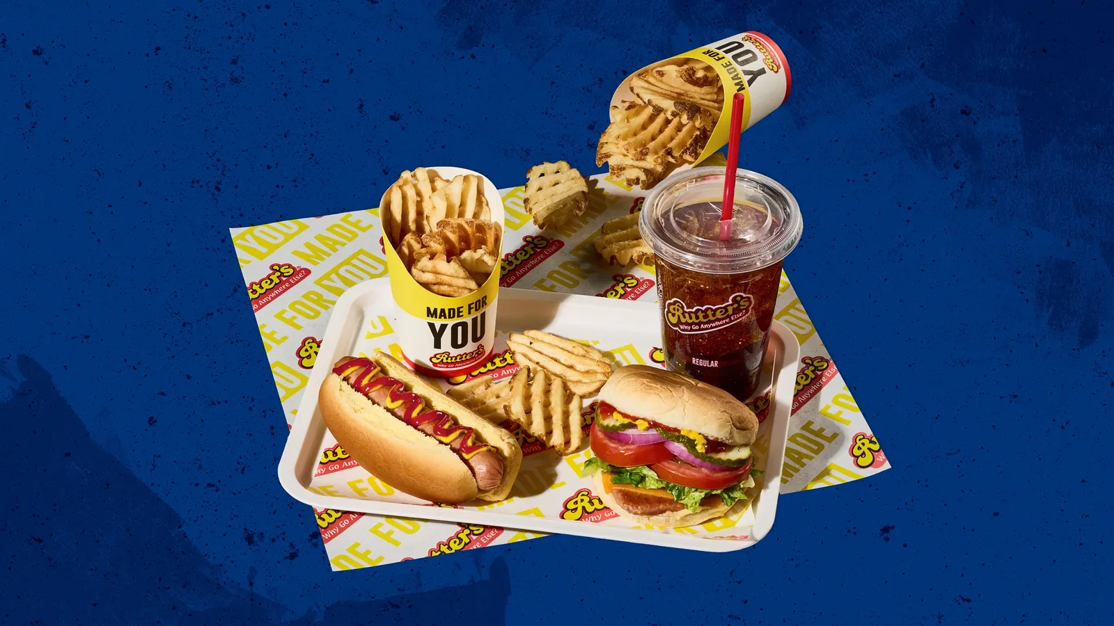A photo of a meal on a tray against a blue background. There are waffle frie, a hot dog, a sandwich and a beverage. The fries cohntainers say made for you and logos on many of the containers and wrappers say Rutter's.