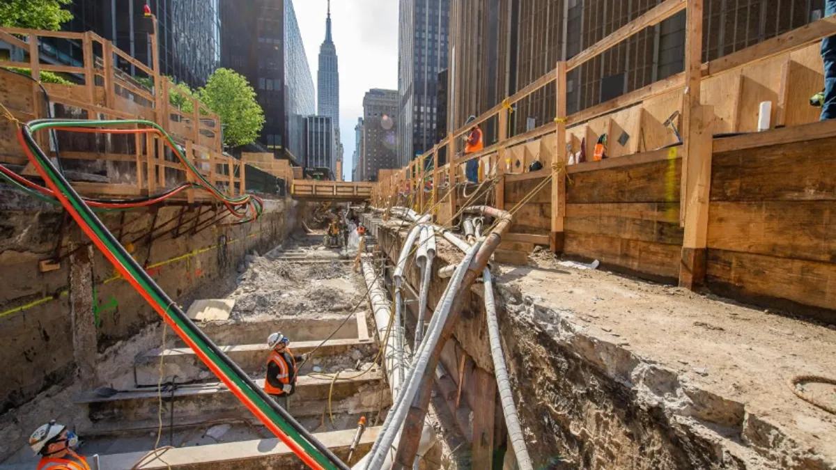 Construction teams work in a massive trench with New York City's skyscrapers in the background.