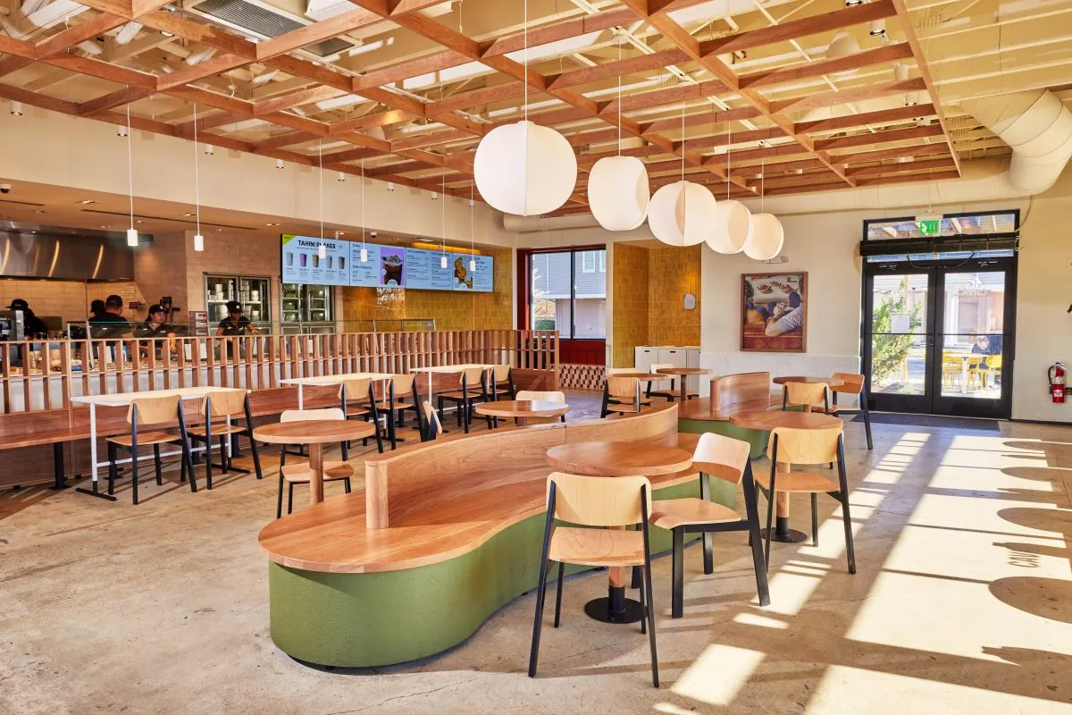 Wooden seats and tables inside a Cava. In the background there is a make line with digital menu boards.