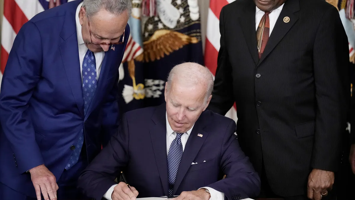 President Joe Biden sits at a desk and signs legislation as Senate Majority Leader Charles Schumer and House Majority Whip James Clyburn stand nearby