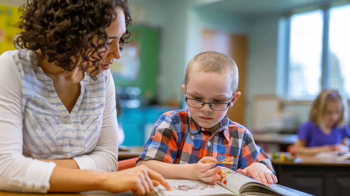 An elementary school student and adult sit at a table in a classroom. They are looking at the inside of a book on the table.
