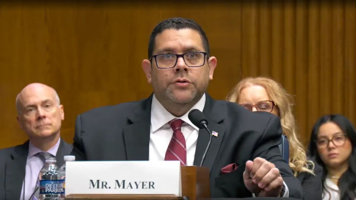 A person in a black suit seated and speaking into a microphone.