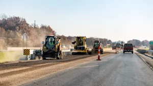 Several construction vehicles on an unpaved section of a highway with cars passing on the adjacent pavement.