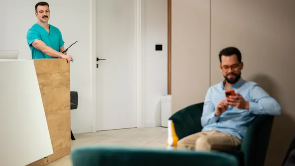 A medical professional standing at a reception desk with a patient waiting.