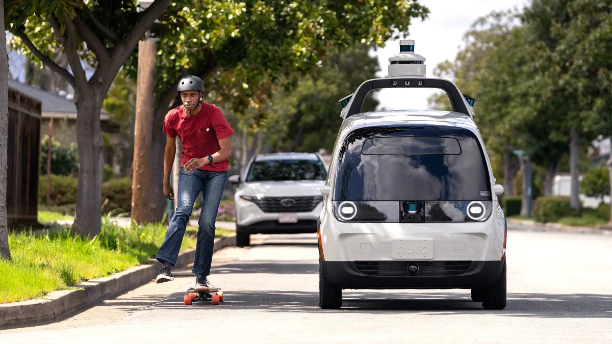 The Nuro R3 autonomous delivery vehicle on the road next to a person riding a skateboard.