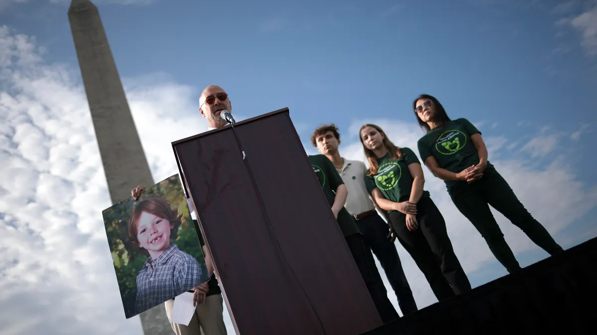 An adult stands behind a podium near the Washington Monument in Washington, D.C. To the adult's right, there is a large photo of a child. People are standing behind  the adult.