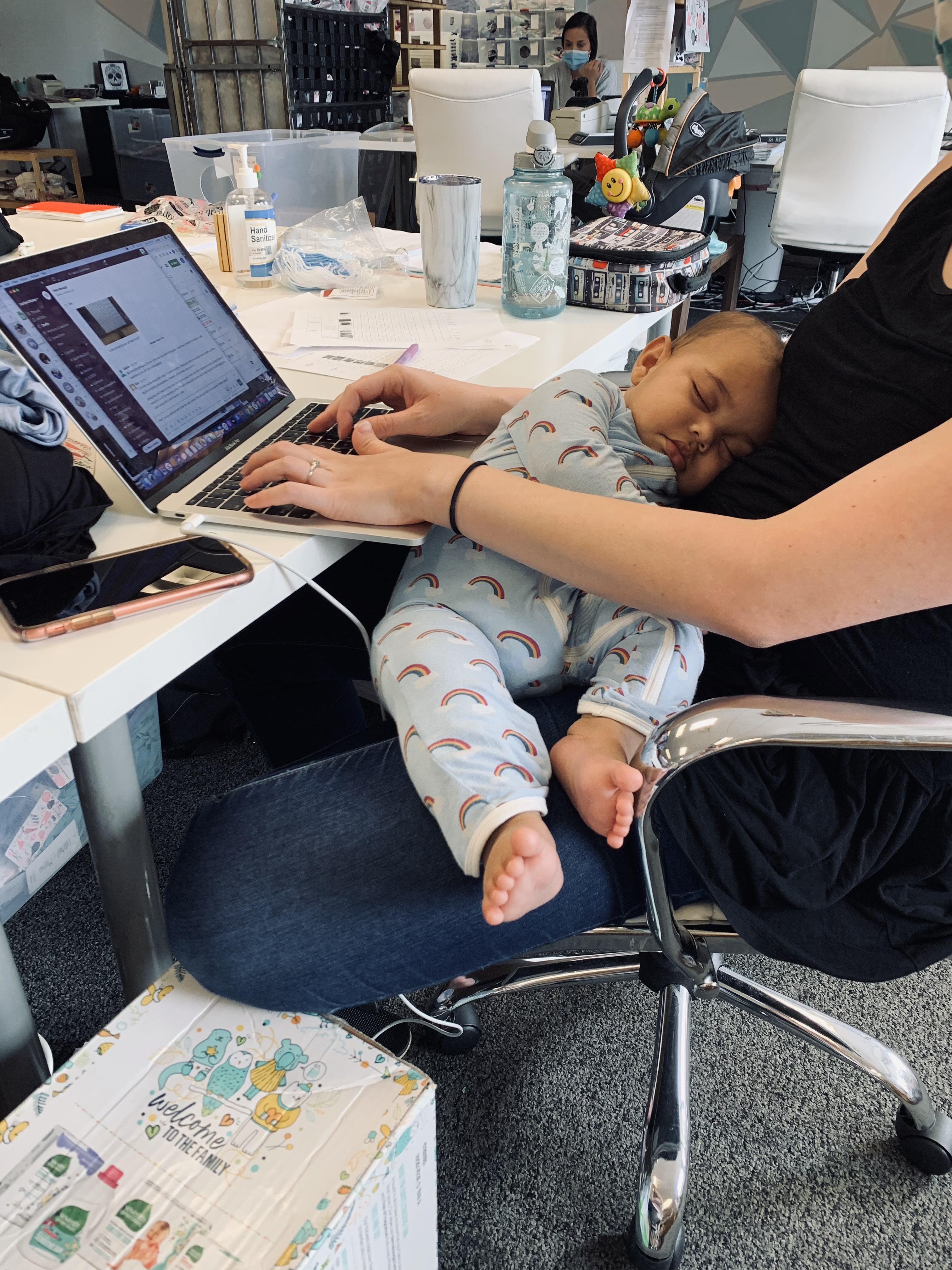 A baby sleeps on its mother's lap as she sits in front of a desk in the office.