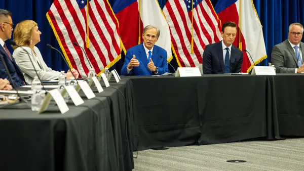 Two people wearing suits sit at a table with U.S. and Texas flags behind them and other people sitting around them.