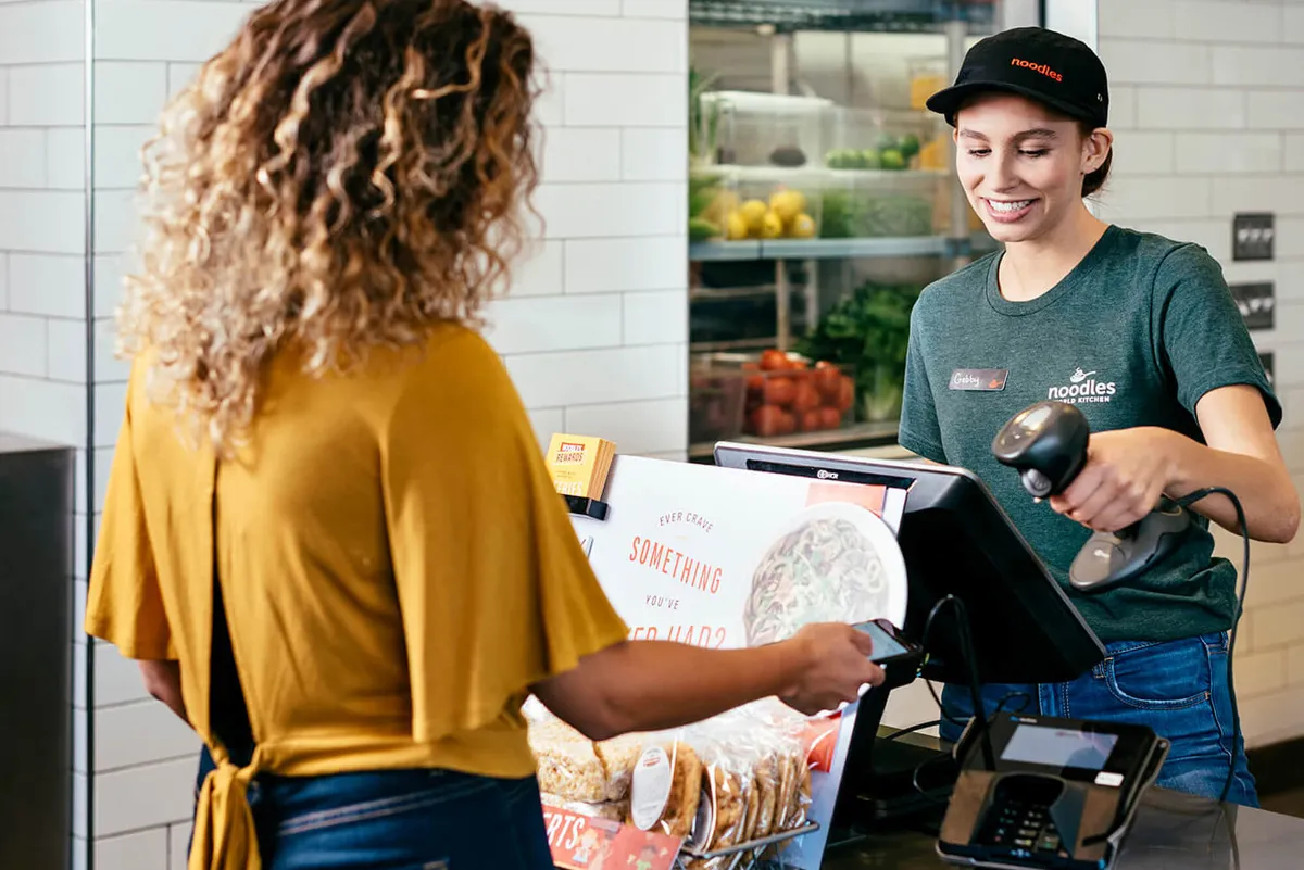 An image of a customer showing a smartphone to a Noodles &amp; Company cashier