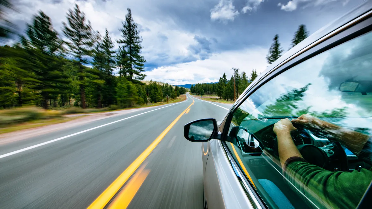 Seen from looking out the left rear window, a car is driving along a two-lane road lined with evergreen trees, leading toward mountains and clouds.