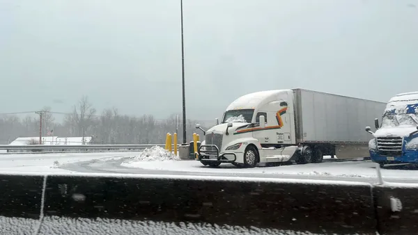 Snow covers windshields of trucks and pavement in a parking area.