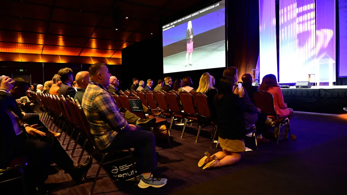 Rows of seated people facing a stage and large screen showing a woman speaking.