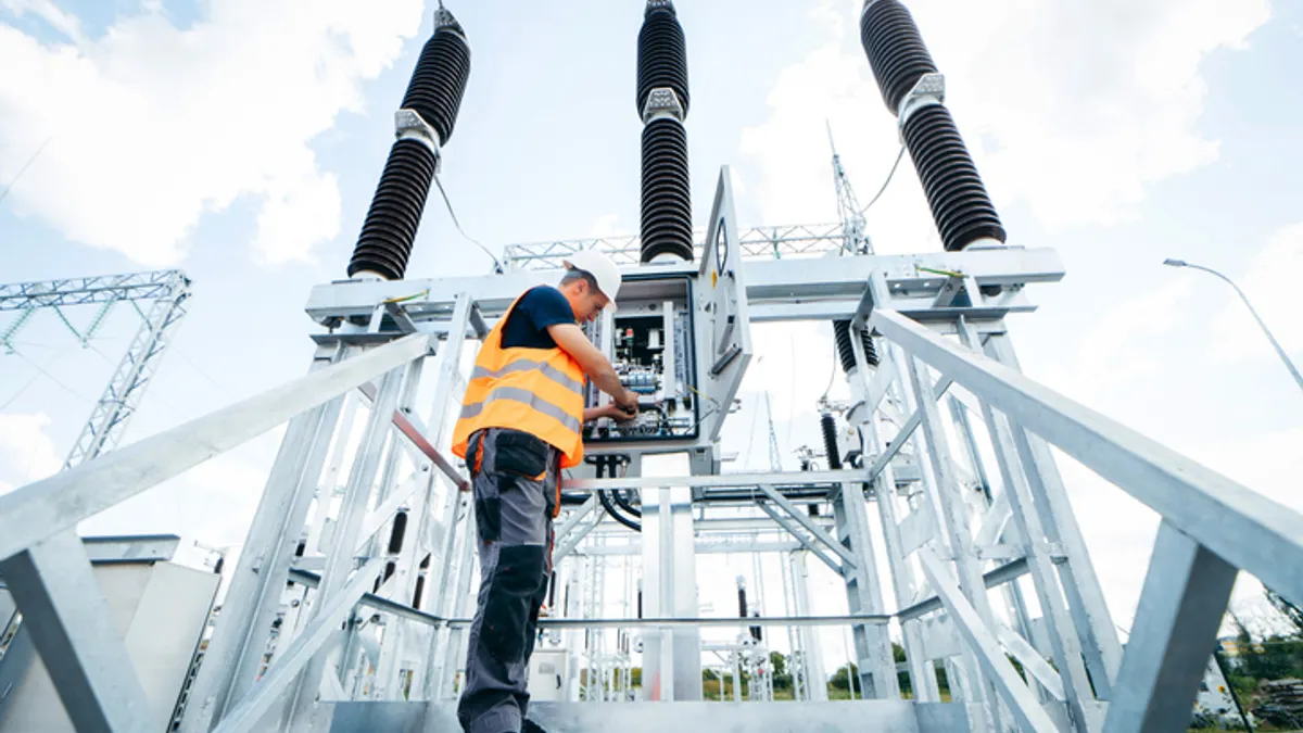 A man wearing an orange vest is positioned on a platform adjacent to a substation, monitoring the surroundings.