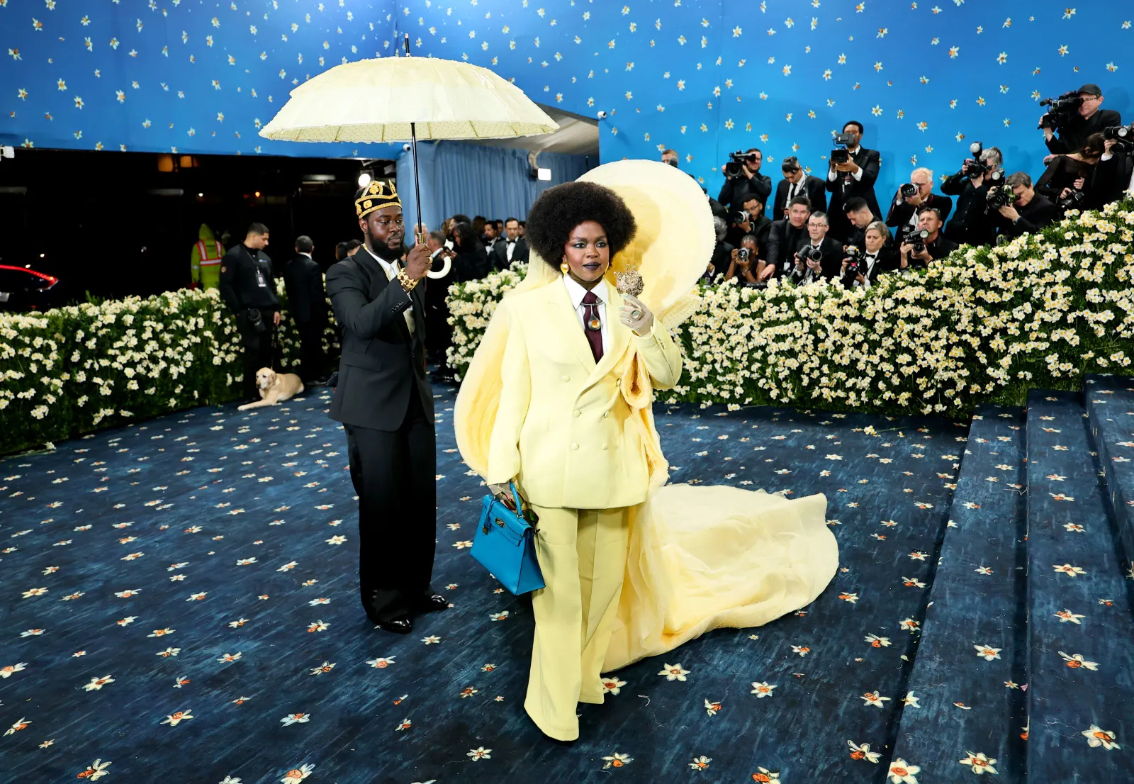 A woman in a yellow suit with a long train holds a blue handbag and stands on a blue carpet. A man behind her holds a yellow umbrella over her head.