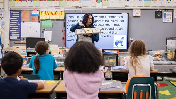 An adult stands at the front of a classroom holding a book. Several students sitting in desks facing the adult are pictured with their backs to the camera.