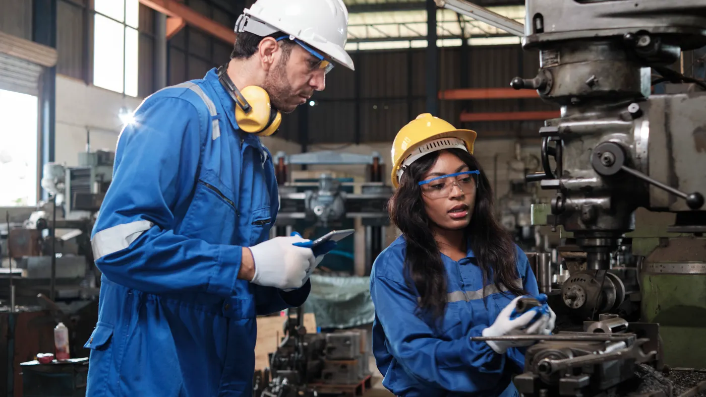 Two industrial workers in protective and safety uniforms and hardhats working with metalwork machines in a manufacturing factory.