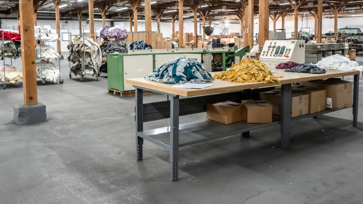 Piles of textiles sorted by color sit on racks and tables in a big warehouse.