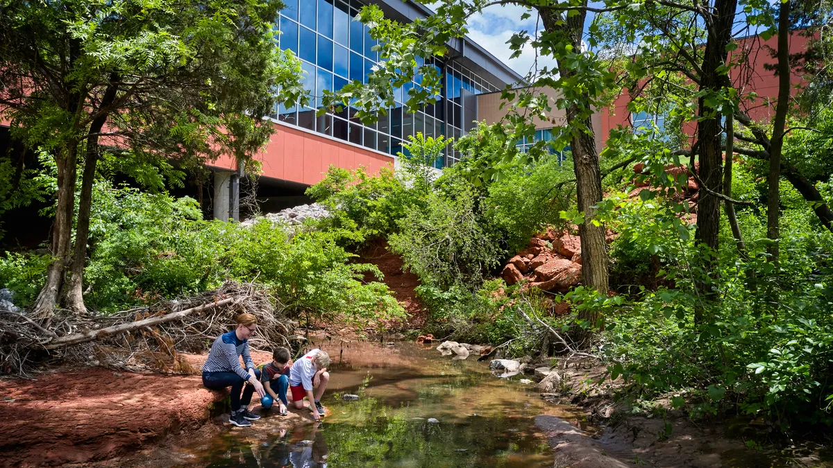 An adult and two student lean down toward a creek surrounded by a school building and vegetation.