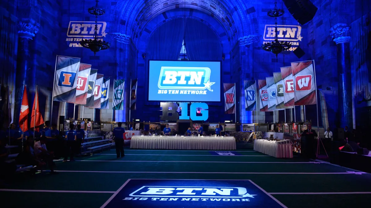 A room is illuminated blue. A table with a white cloth sits in the center with university flags hung to either side, with a large TV over head displaying the Big Ten logo.