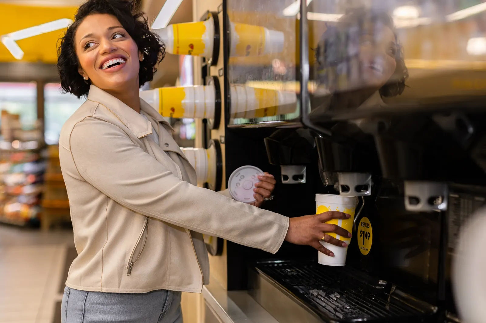 A photo of a person pouring a beverage from a large dispenser in a store into a white and yellow disposable cup. the person has a lid for the cup in the other hand.