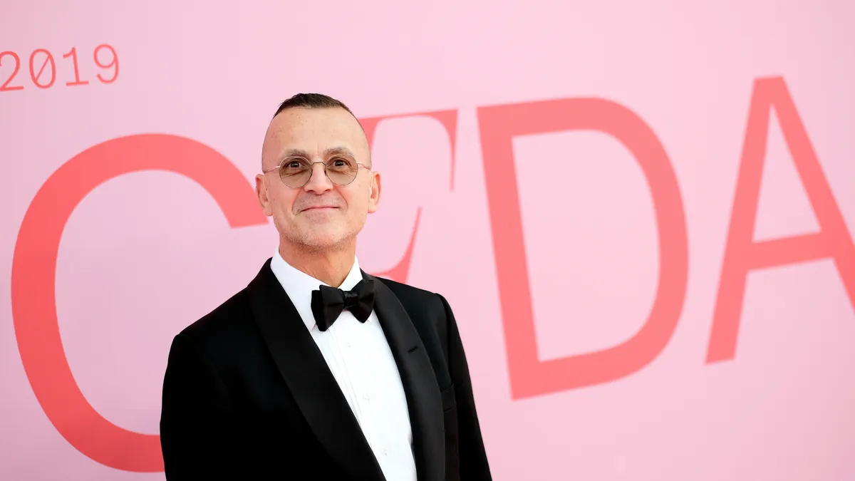 Council of Fashion Designers of America CEO Steven Kolb wears a tuxedo and round sunglasses and stands in front of a pink CFDA sign.