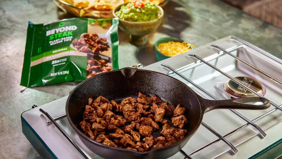 A small cast iron skillet with Beyond Steak bites inside sits on a gas-powered hot plate with the product package and a dish of tortilla chips with dips is in the background.