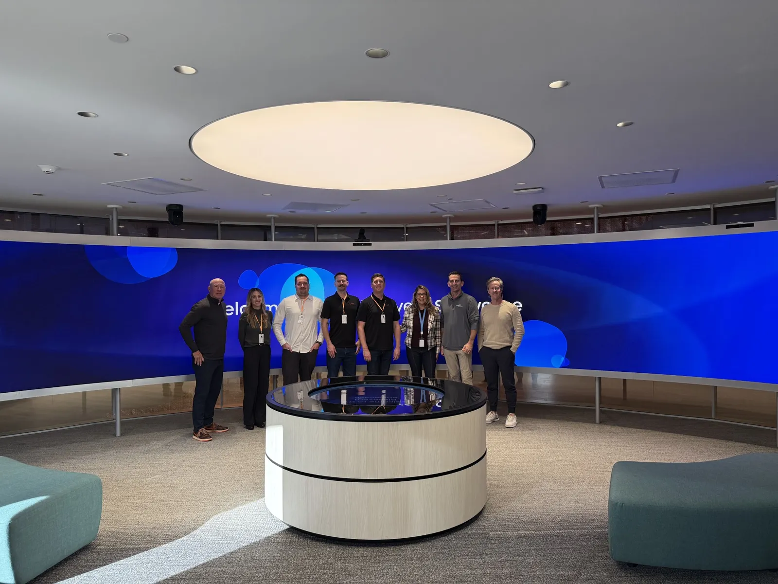 Eight people stand in front of a long blue monitor and behind a white circular table.
